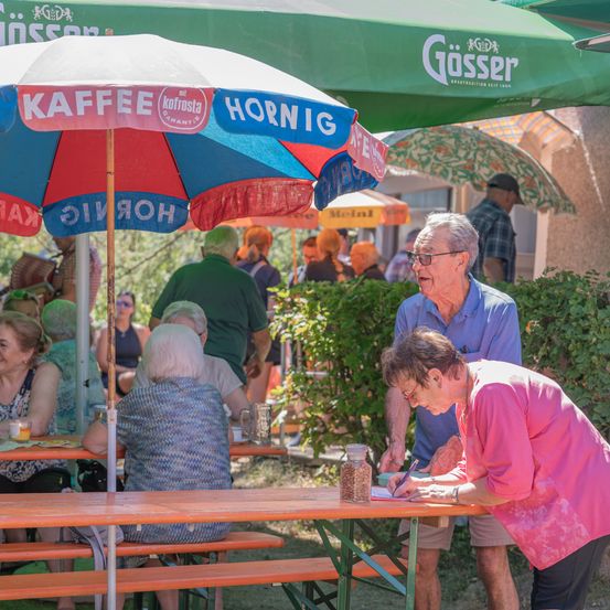 Eine ältere Frau schreibt auf einem Stück Papier an einem Tisch in einem Restaurant im Freien, während ein älterer Mann zusieht. Der Tisch ist mit einem Schirm bedeckt, auf dem Kaffee Hornig steht.