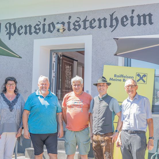Fünf Personen, lässig gekleidet, stehen vor einem Gebäude mit dem Schild 'Pensionörstenheim'. Der Mann in der Mitte trägt einen Hut und eine Weste. Hinter ihnen ist ein gelbes Banner mit dem Text 'Raiffeisen Meine Bank' zu sehen.