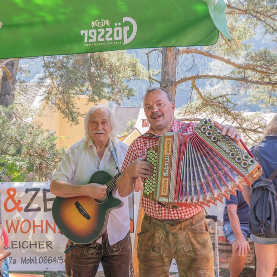 Zwei Männer in traditioneller bayerischer Kleidung, einer spielt Akkordeon und der andere Gitarre, stehen unter einem grünen Vordach mit einem Banner im Hintergrund.