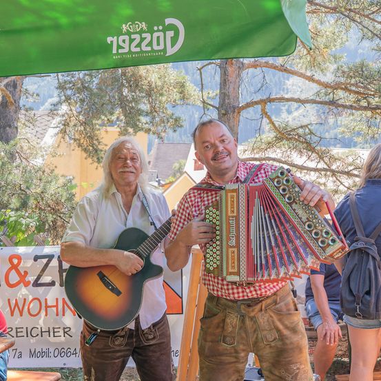 Zwei Männer in traditioneller bayerischer Kleidung, einer spielt Gitarre und der andere hält ein Akkordeon, stehen unter einem grünen Vordach mit einem Banner im Hintergrund.