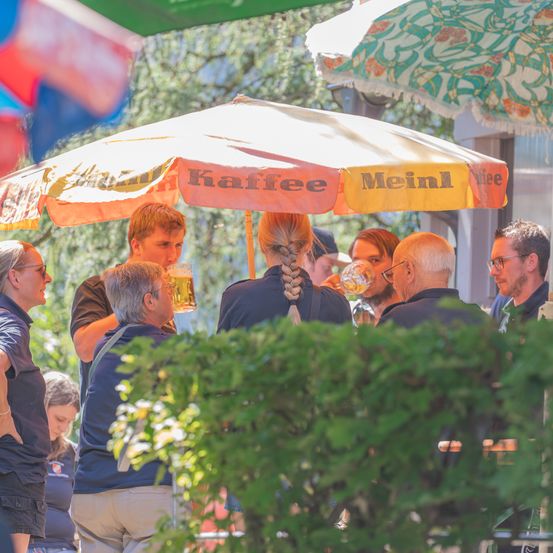 Eine Gruppe von Menschen versammelt sich unter einem Regenschirm in einem Biergarten, einige trinken Bier, während andere in Gespräche vertieft sind.