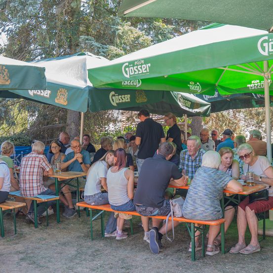 Eine Gruppe von Menschen sitzt unter grünen Schirmen in einem Biergarten im Freien, einige trinken und andere unterhalten sich.