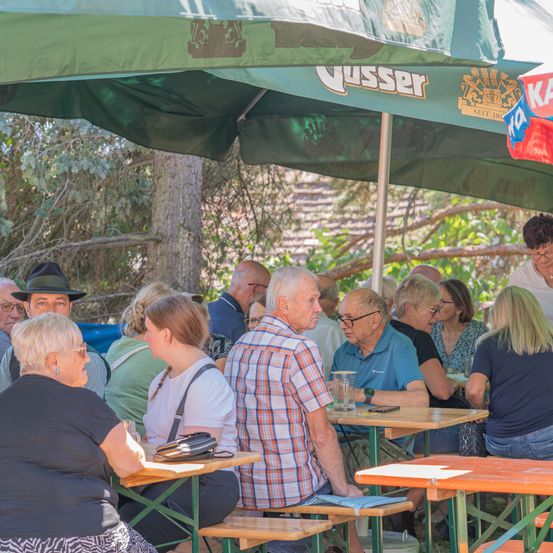 Eine Gruppe älterer Erwachsener sitzt unter einem grünen Schirm in einem Biergarten, einige unterhalten sich und andere schauen sich um. Die Tische sind mit Gläsern bestückt und es gibt Bäume im Hintergrund.