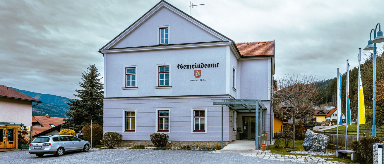 Ein weißes Gebäude mit roten Verzierungen und einem roten Dach trägt den Schriftzug Gemeindeamt. Es ist von Bäumen und Pflanzen umgeben. Ein Auto ist davor geparkt.