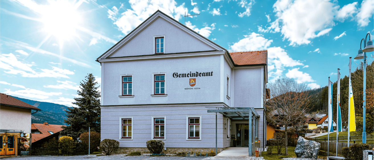 Ein weißes Gebäude mit roten Akzenten und einem roten Dach, über dem Eingang steht 'Gemeindeamt'. Das Gebäude ist von Grünpflanzen umgeben.