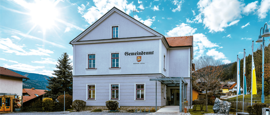Ein weißes Gebäude mit roten Akzenten und einem roten Dach, über dem Eingang steht 'Gemeindeamt'. Das Gebäude ist von Grünpflanzen umgeben.