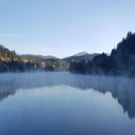 Ein ruhiger See mit nebliger Atmosphäre und Blick auf die umliegenden Berge und Bäume unter einem klaren blauen Himmel.