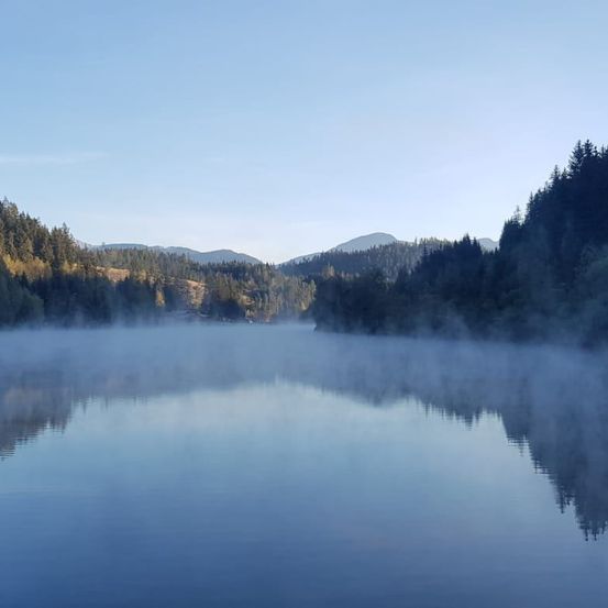 Ein ruhiger See mit nebliger Atmosphäre und Blick auf die umliegenden Berge und Bäume unter einem klaren blauen Himmel.