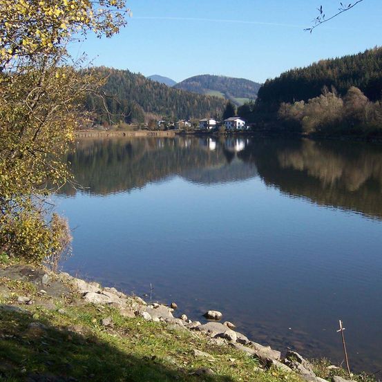 Ein ruhiger See spiegelt den blauen Himmel und Bäume wider, mit einem entfernten Haus und Bergen.