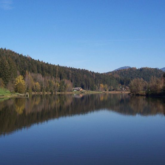 Ein ruhiger See umgeben von üppigen Wäldern und Bergen unter einem klaren blauen Himmel. Spiegelungen von Bäumen und Gebäuden spiegeln sich auf der ruhigen Wasseroberfläche.