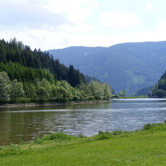 Ein ruhiger See mit sanften Wellen, umgeben von üppigen grünen Bäumen und entfernten Bergen unter einem blauen Himmel mit Wolken.