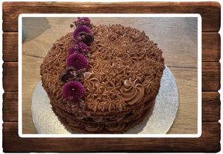 Ein Schokoladenkuchen, bedeckt mit Schokoladenglasur, ist mit lila Blumen und Schokoladenherzen verziert. Er steht auf einem silbernen Teller auf einer Holzoberfläche.