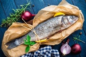 Ein ganzer Fisch in braunes Papier gewickelt und mit einem Faden gebunden, mit Zitronenscheiben, Kräutern und Zwiebeln auf einem blauen Tisch.