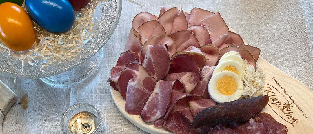 Ein Holzbrett mit Fleisch, Eiern und Bohnen. Eine Glasschale mit bunten Eiern. Ein Teller mit Brot, Bohnen und einer Gabel. Eine Kerze in einem Glashalter. Ein Holzschneidebrett auf dem Tisch.