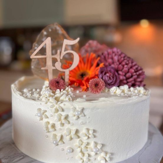 Ein weißer Kuchen mit der Nummer 45 oben, verziert mit verschiedenen Blumen und Perlen. Der Kuchen hat einen Marmorkuchenständer.