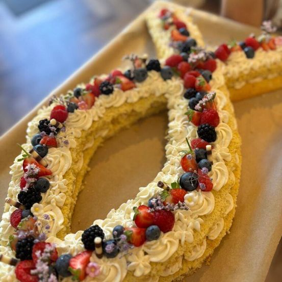 Ein gelber Kuchen in Form der Zahl 8 ist mit weißer Glasur und einer Vielzahl von Beeren und Blumen oben dekoriert.