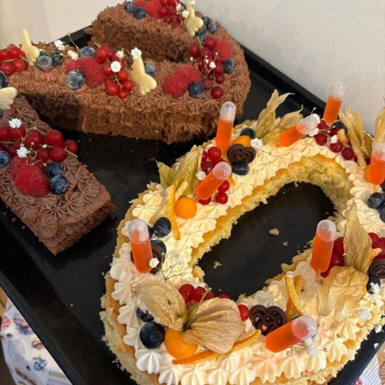 Drei Schokoladenkuchen in Form der Zahl 0, verziert mit Beeren, Blumen und orangefarbenen Geltuben, stehen auf einem schwarzen Tablett. Ein Kuchen ist vollständig mit Schokolade überzogen.