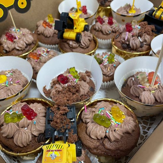 Eine Schachtel Schokoladen-Cupcakes mit verschiedenen Toppings wie Schokolade, Streuseln und Gummibärchen. Einige Cupcakes haben einen Spielzeugbagger obenauf.