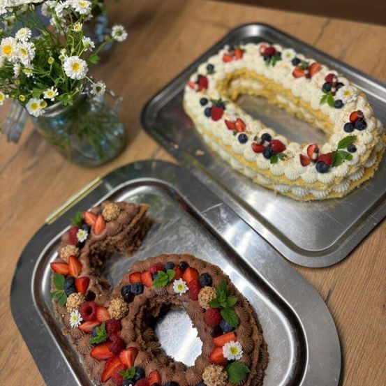 Zwei Schokoladen- und Sahnetorten in Form der Zahl 6 auf einem Tablett, dekoriert mit frischen Früchten und Blumen. Ein Glas mit weißen Blumen steht auf einem Holztisch.