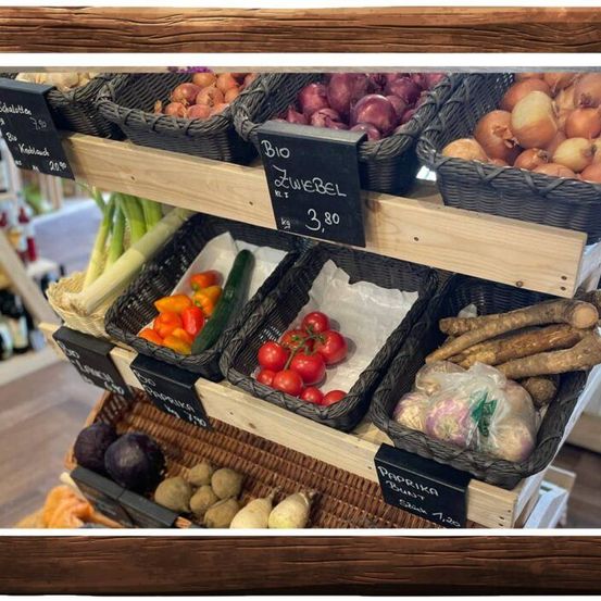 Ein Holzregal mit Körben verschiedener Gemüsesorten wie Zwiebeln, Tomaten, Paprika und anderen. Auf jedem Korb befinden sich Preisschilder.