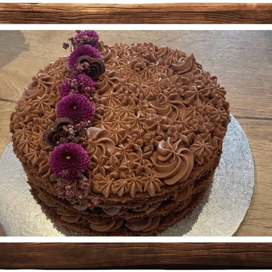 Ein Schokoladenkuchen mit einer dicken Schicht Schokoladenglasur, verziert mit lila Blumen und herzförmigen Schokoladen, auf einem Silberteller auf einem Holztisch.