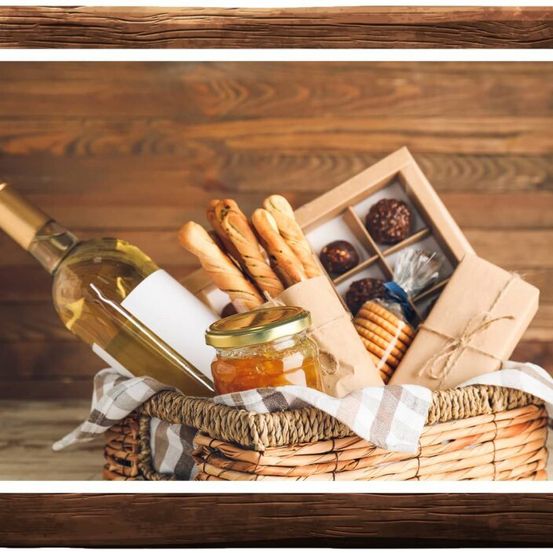Ein Korb aus Weiden mit einer Flasche Weißwein, einem Glas Marmelade, Brot und einer Schachtel Pralinen vor einem hölzernen Hintergrund.