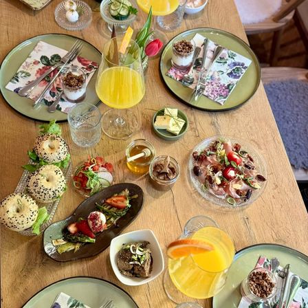 Ein hölzerner Tisch ist mit verschiedenen Gerichten und Getränken gedeckt. Bagels, Salate und kleine Teller mit Obst und Gemüse sind arrangiert. Es gibt Gläser mit Orangensaft, Wasser und eine Desserttasse. Die Umgebung umfasst Blumen und Besteck.