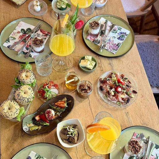 Ein hölzerner Tisch ist mit verschiedenen Gerichten und Getränken gedeckt. Bagels, Salate und kleine Teller mit Obst und Gemüse sind arrangiert. Es gibt Gläser mit Orangensaft, Wasser und eine Desserttasse. Die Umgebung umfasst Blumen und Besteck.