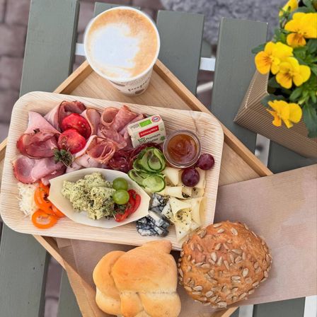 Ein Holztablett hält eine vielfältige Auswahl an Fleisch, Käse, Gemüse und Brot. Ein Kaffeebecher mit Schaumkünstchen steht neben dem Tablett. Ein Vasenstrauß mit gelben Blumen befindet sich rechts.