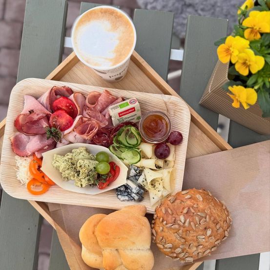 Ein Holztablett hält eine vielfältige Auswahl an Fleisch, Käse, Gemüse und Brot. Ein Kaffeebecher mit Schaumkünstchen steht neben dem Tablett. Ein Vasenstrauß mit gelben Blumen befindet sich rechts.
