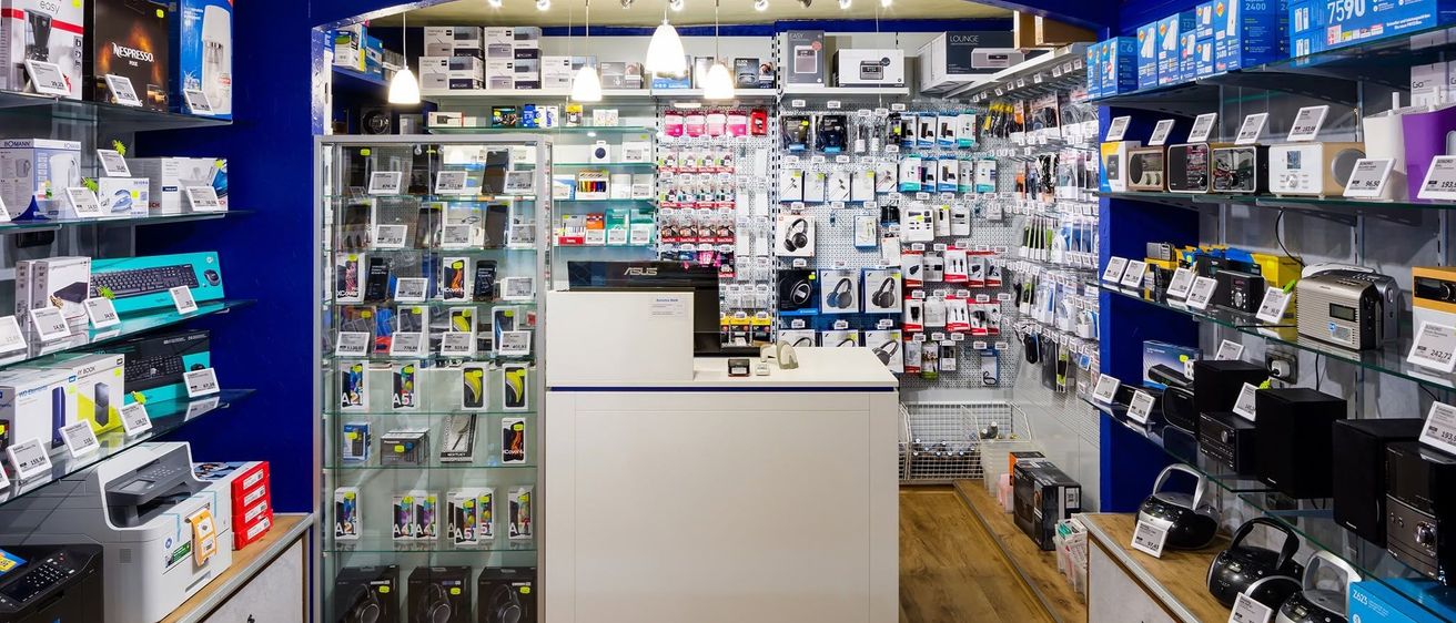 Ein blauer und weißer Elektronikladen mit Holzboden. Eine Theke zeigt verschiedene Produkte. Regale halten zahlreiche Artikel. Ein Regal enthält Kisten und ein anderes zeigt Kameras. Über der Theke hängen Lampen.