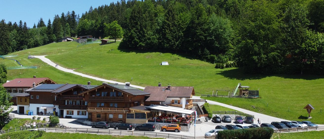 Aerial view of a rustic house with a brown roof surrounded by a green meadow and tall trees. There are cars parked in front of the building, and a playground is visible in the distance.