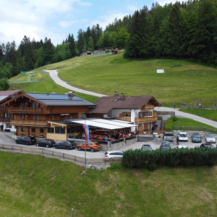 Eine Draufsicht auf ein Chalet mit Solarmodulen, umgeben von einem üppigen grünen Hang, mit mehreren geparkten Autos davor und einem Pfad, der den Hügel hinaufführt.