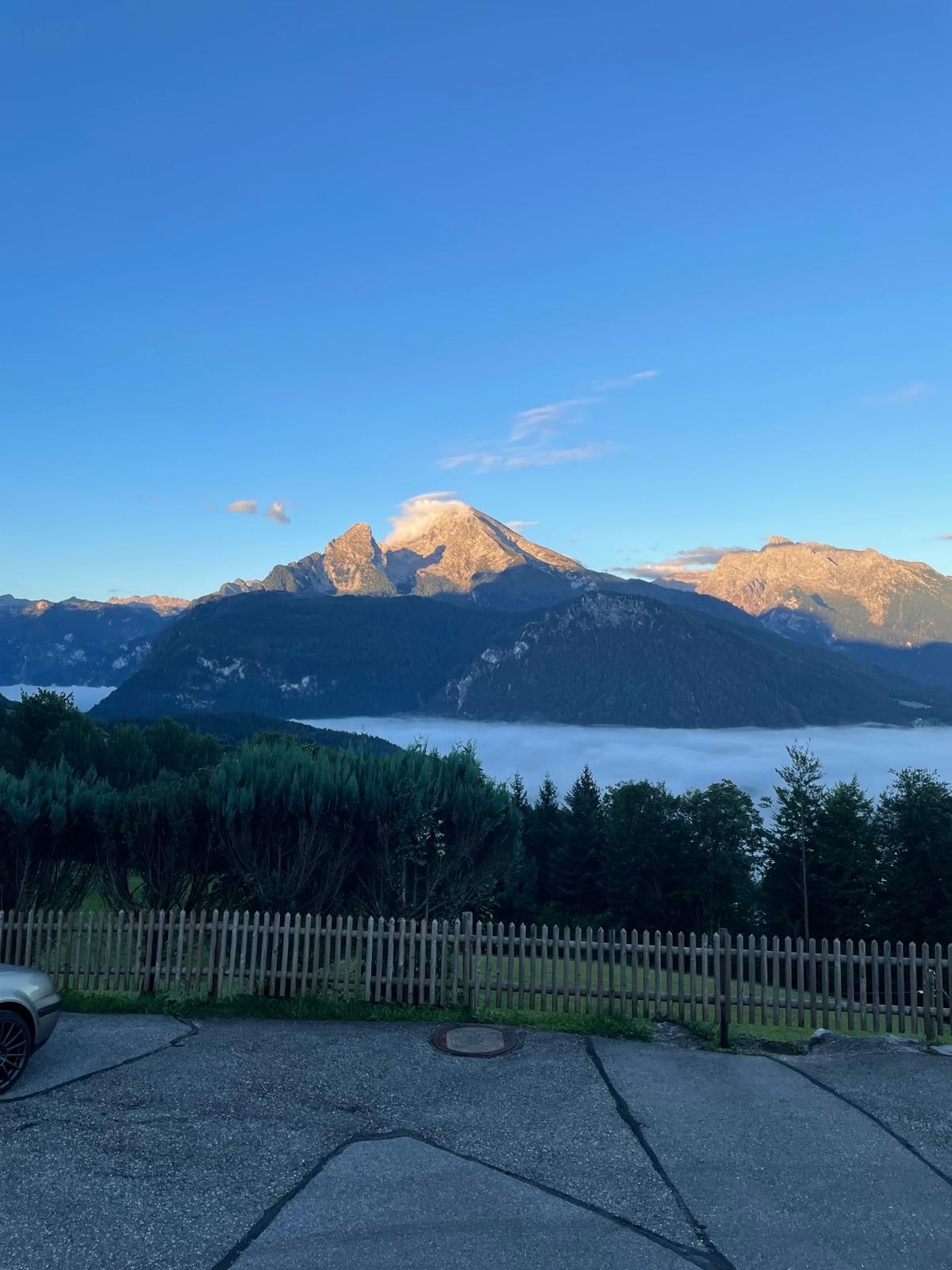 Eine malerische Aussicht auf Berge mit einem See darunter, unter einem blauen Himmel. Ein Auto ist in der Nähe eines Holzzauns geparkt.