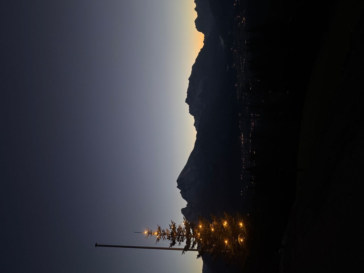 Ein kleiner Weihnachtsbaum mit Lichtern steht vor einer Bergsilhouette unter einem dunklen Himmel.