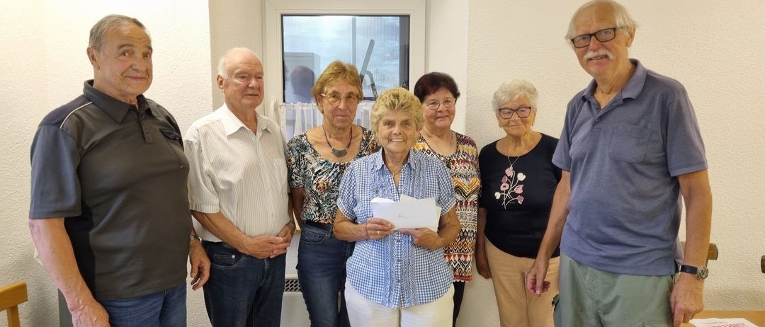 Eine Gruppe älterer Menschen steht in einem Raum, eine Frau hält ein Stück Papier.
