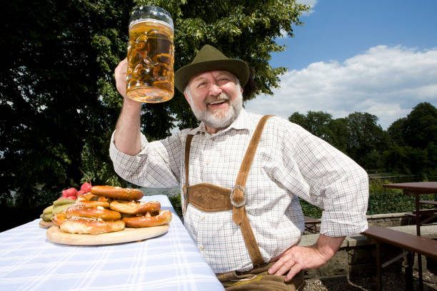 Ein Mann in traditioneller bayerischer Kleidung hält einen großen Bierkrug, lächelt und hat Brezen und andere Speisen auf einem Tisch hinter sich.