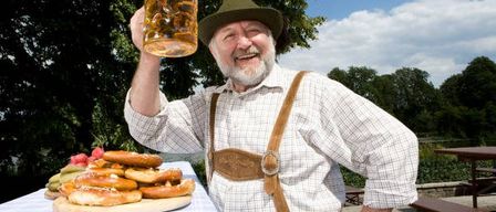 Ein Mann in traditioneller bayerischer Kleidung hält einen großen Bierkrug, lächelt und hat Brezen und andere Speisen auf einem Tisch hinter sich.