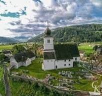 Ein Luftbild eines kleinen Dorfes mit einer weißen Kirche und einem Friedhof im Vordergrund, umgeben von grünen Feldern und Bergen im Hintergrund.