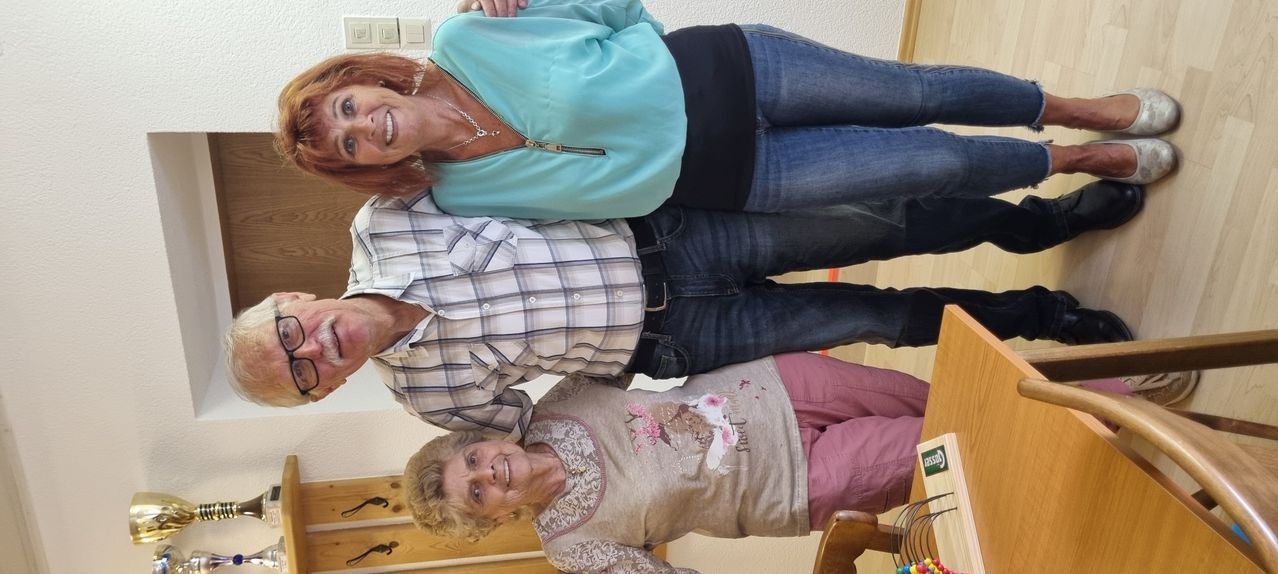 Drei ältere Menschen lächeln für ein Foto, stehen zusammen. Die Frau in der Mitte trägt ein grünes Oberteil. Der Mann und die Frau auf beiden Seiten tragen Freizeitkleidung.