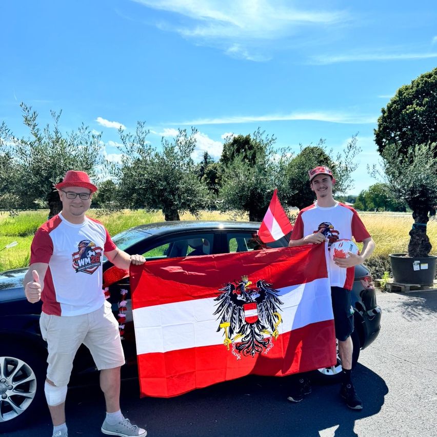 Two men hold an Austrian flag in front of a car in a sunny outdoor setting with trees in the background.