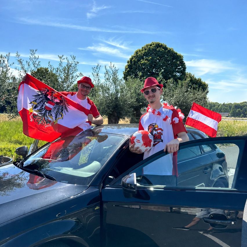 Two men in red hats stand beside a car with a flag in hand. One holds a soccer ball, and both wave flags. Behind them is a field with trees.