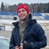 Ein Mann posiert mit einem silbernen Sportwagen auf einer Rennstrecke, mit Red Bull-Markenzeichen, vor einer leeren Tribüne.
