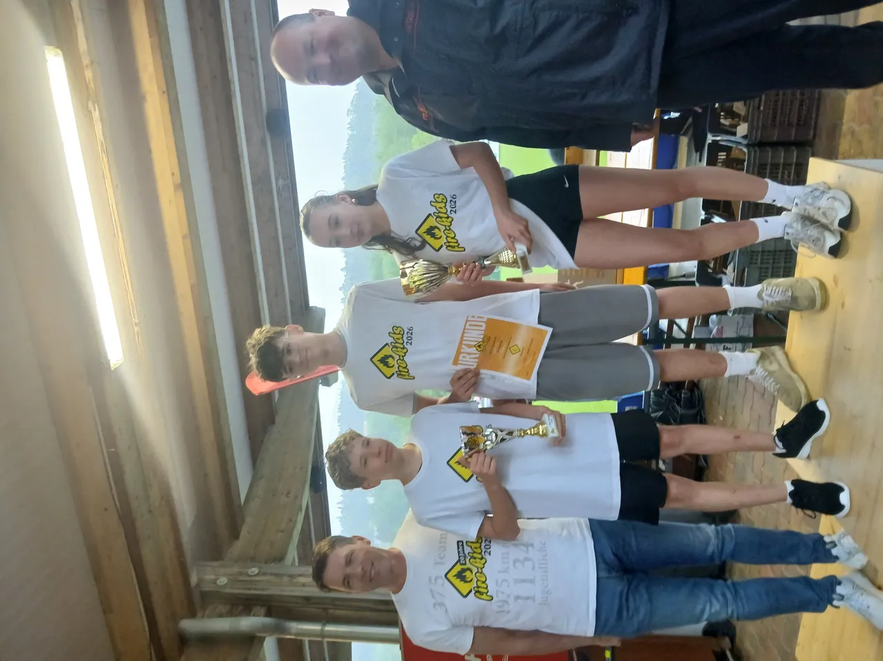 Fünf junge Personen, alle in weißen T-Shirts, posieren mit Pokalen für ein Foto. Der Hintergrund ist eine Holzkonstruktion mit einem Glasfenster.