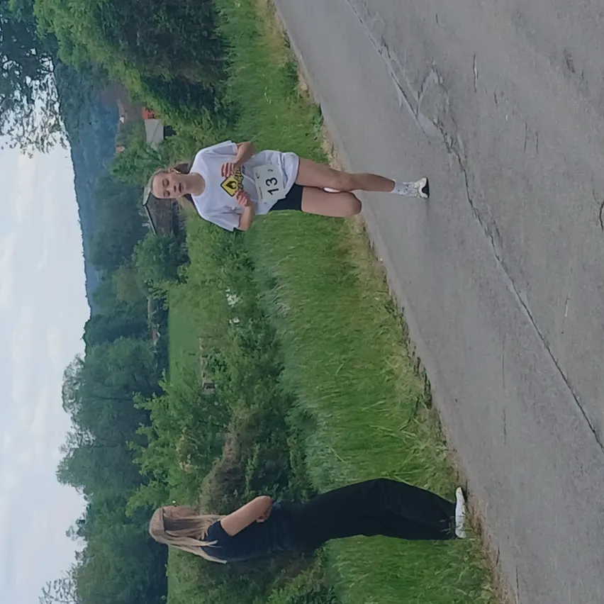 Eine Läuferin mit der Nummer 13 auf dem Shirt läuft auf einer Straße mit einem Grasrand. Eine andere Frau steht auf dem Gras auf der linken Seite.