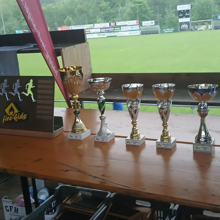 Ein Holztisch mit fünf Trophäen, eine mit einem fire-kids-Logo. Im Hintergrund ist ein roter Banner und ein Sportfeld zu sehen.