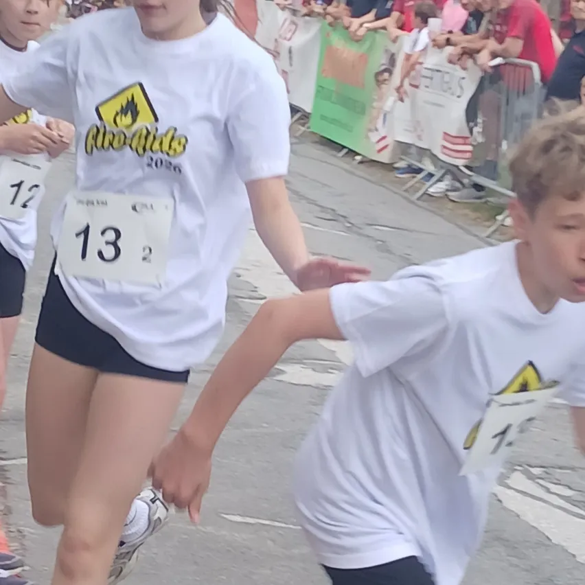 Ein junges Mädchen in einem weißen Shirt mit Nummer 13 läuft in einem Rennen und führt einen Jungen mit Nummer 1 in der Nähe. Zuschauer beobachten hinter einer Barriere.