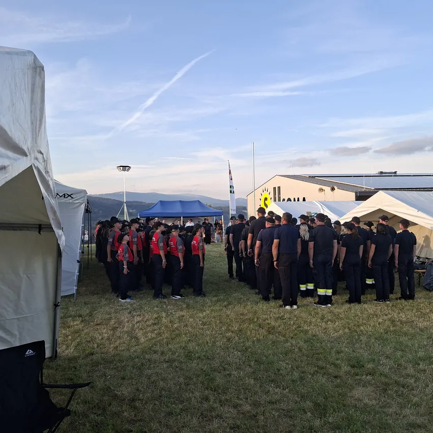 Eine Gruppe von Menschen in Uniform steht in einer Reihe auf einem Rasenfeld, mit Zelten und einem Gebäude im Hintergrund.