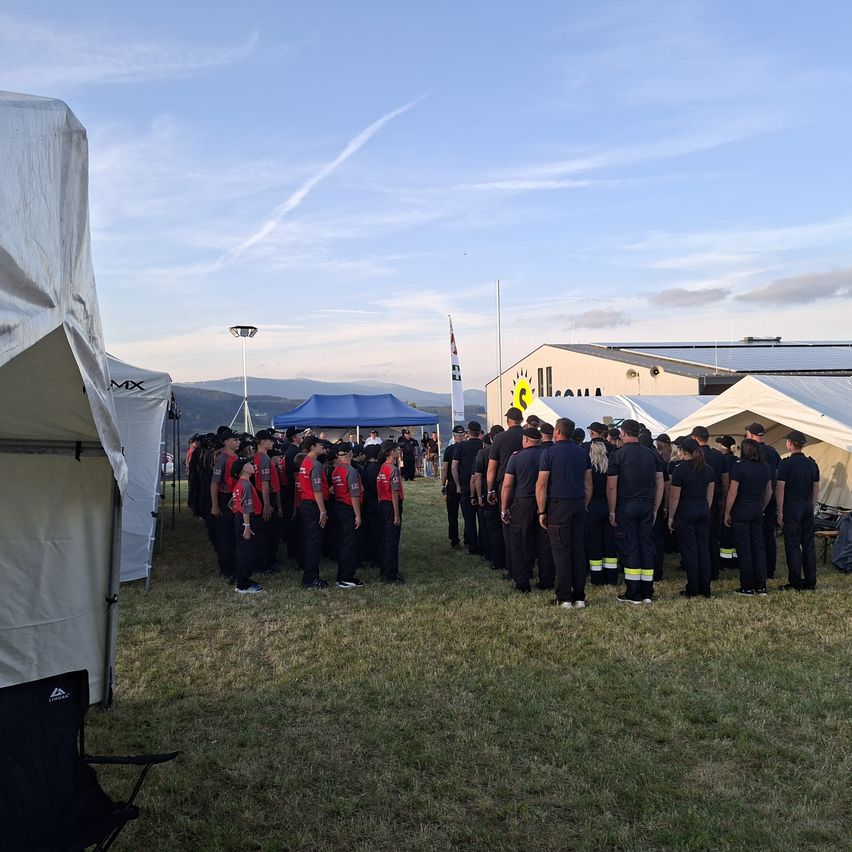 Eine Gruppe von Menschen in Uniform steht in einer Reihe auf einem Rasenfeld, mit Zelten und einem Gebäude im Hintergrund.