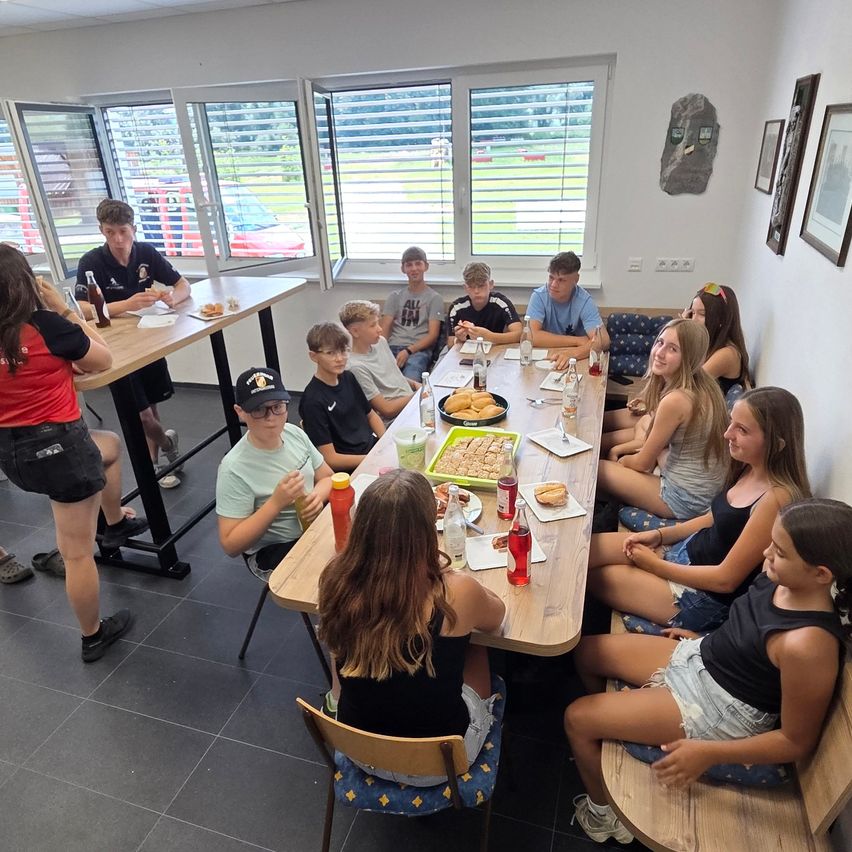 Eine Gruppe von Teenagern und jungen Erwachsenen sitzt um einen langen Tisch in einem Raum mit großen Fenstern und genießt Essen und Getränke.